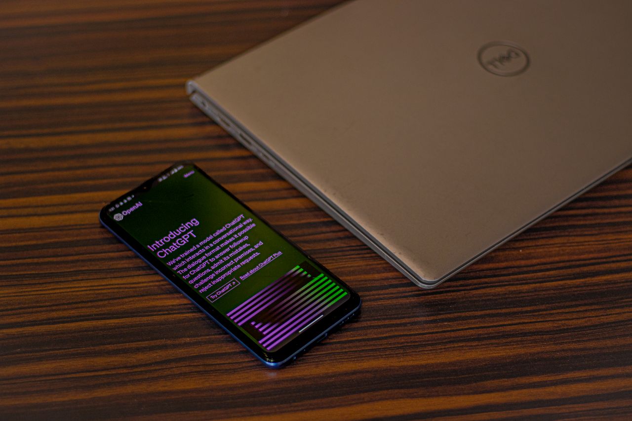 A smartphone displaying ChatGPT app beside a closed laptop on a wooden table.